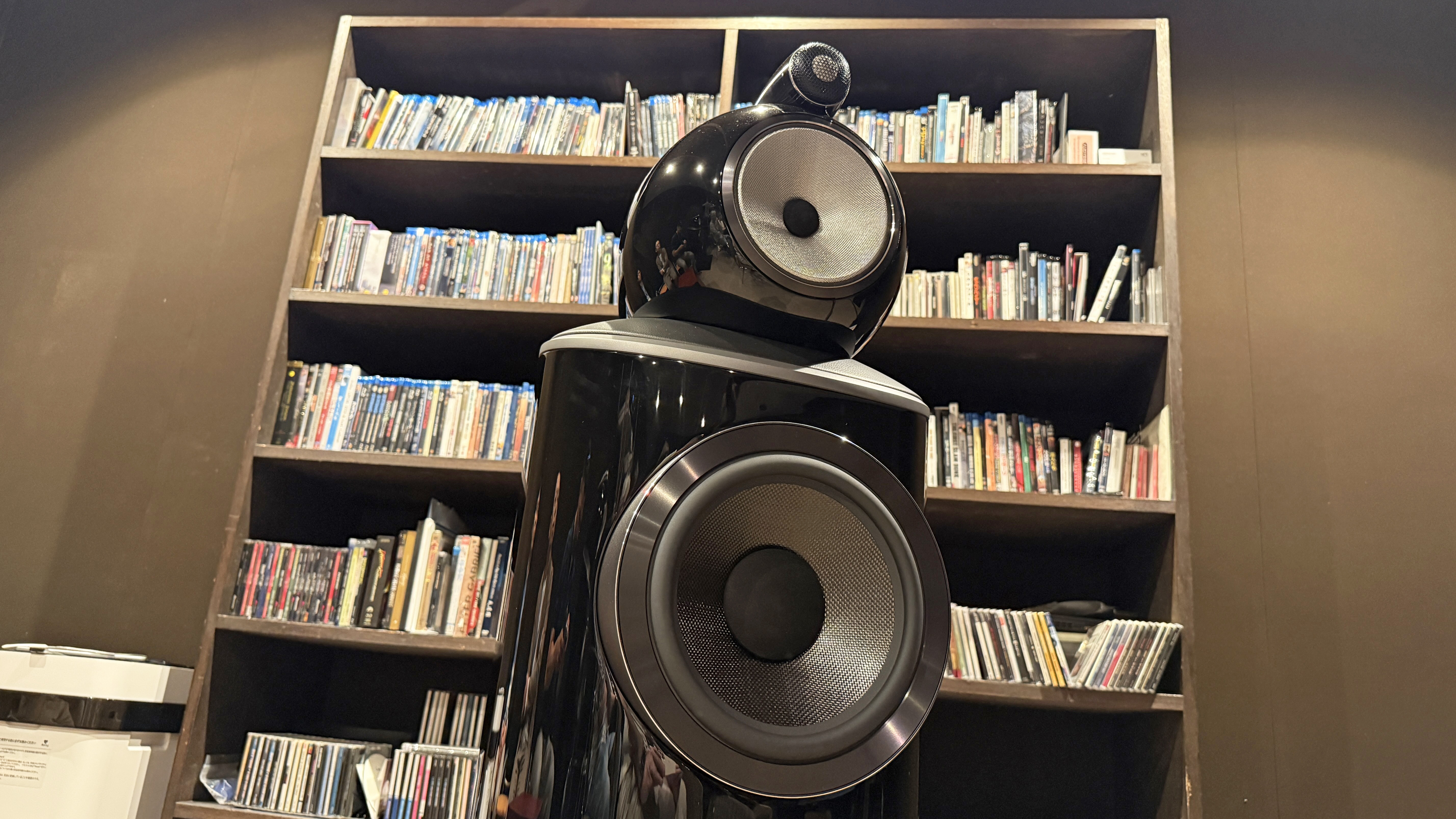 The interior of a home theater listening room with a Bowers &amp; Wilkins 801 D4 speaker in front of a shelf full of Blu-rays and CDs