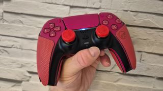 A PS5 DualSense controller sporting Kontrol Freek thumbstick grips and controller grips being held in a man's hand in front of a white brick wall