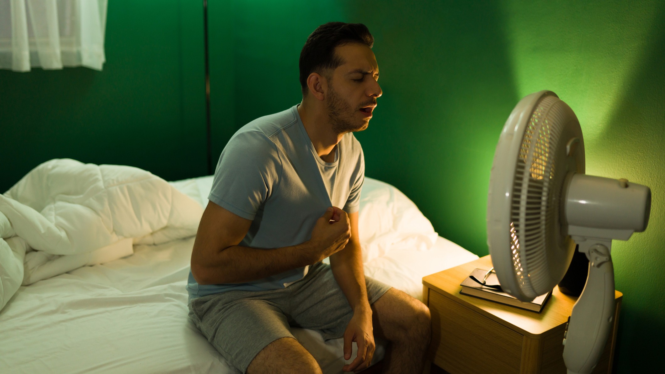 Sweaty man sitting on bed at night, facing electric fan