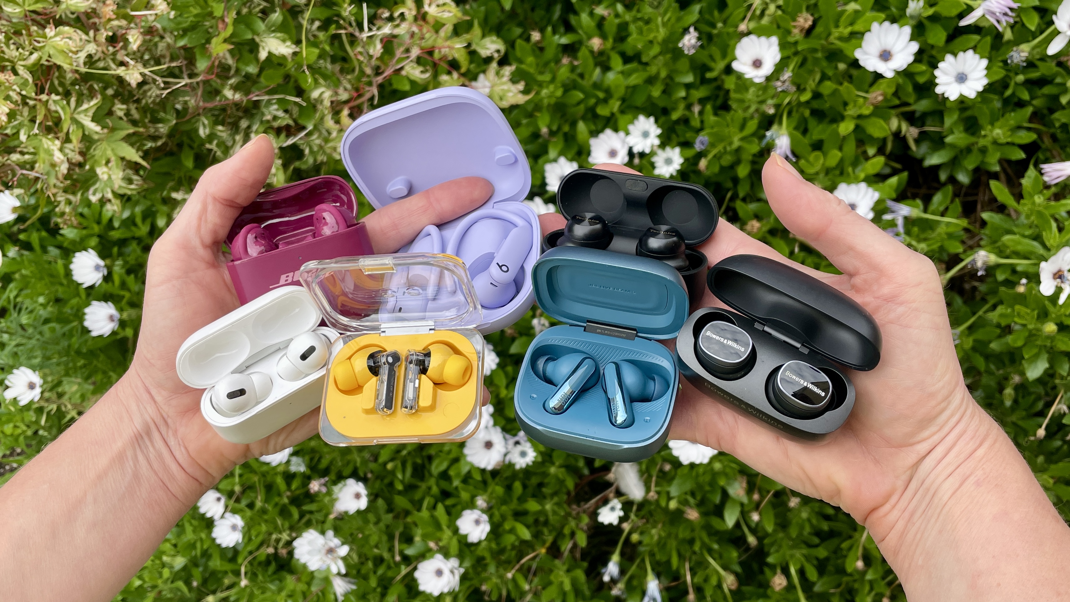 A selection of earbuds (JBL, Bose, Beats, Bowers &amp; Wilkins, Technics and Apple AirPods) held in two hands, in front of a flower bed