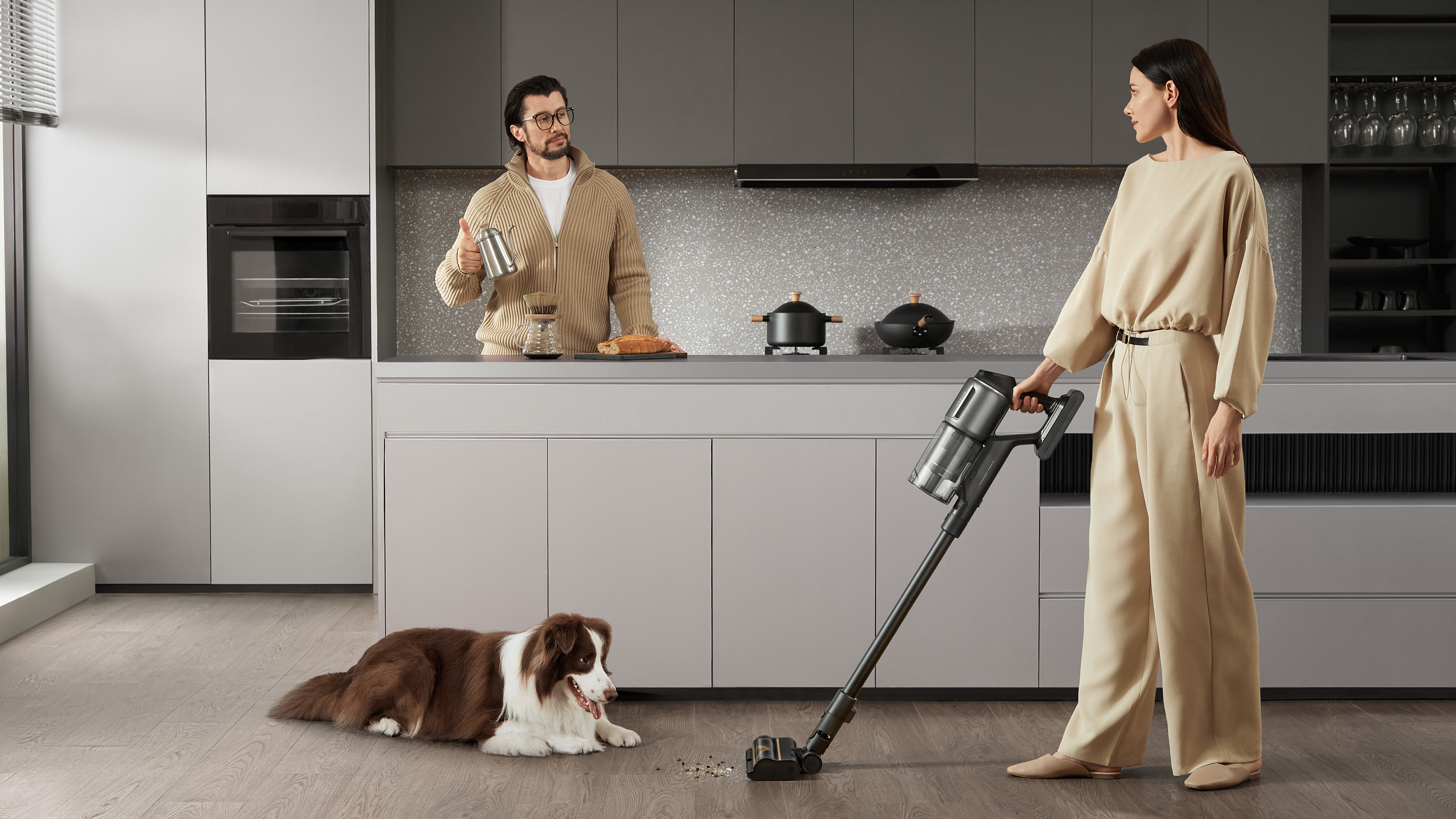 One person makes coffee while another uses a Dreame Cordless Stick Vacuum. A dog looks on.