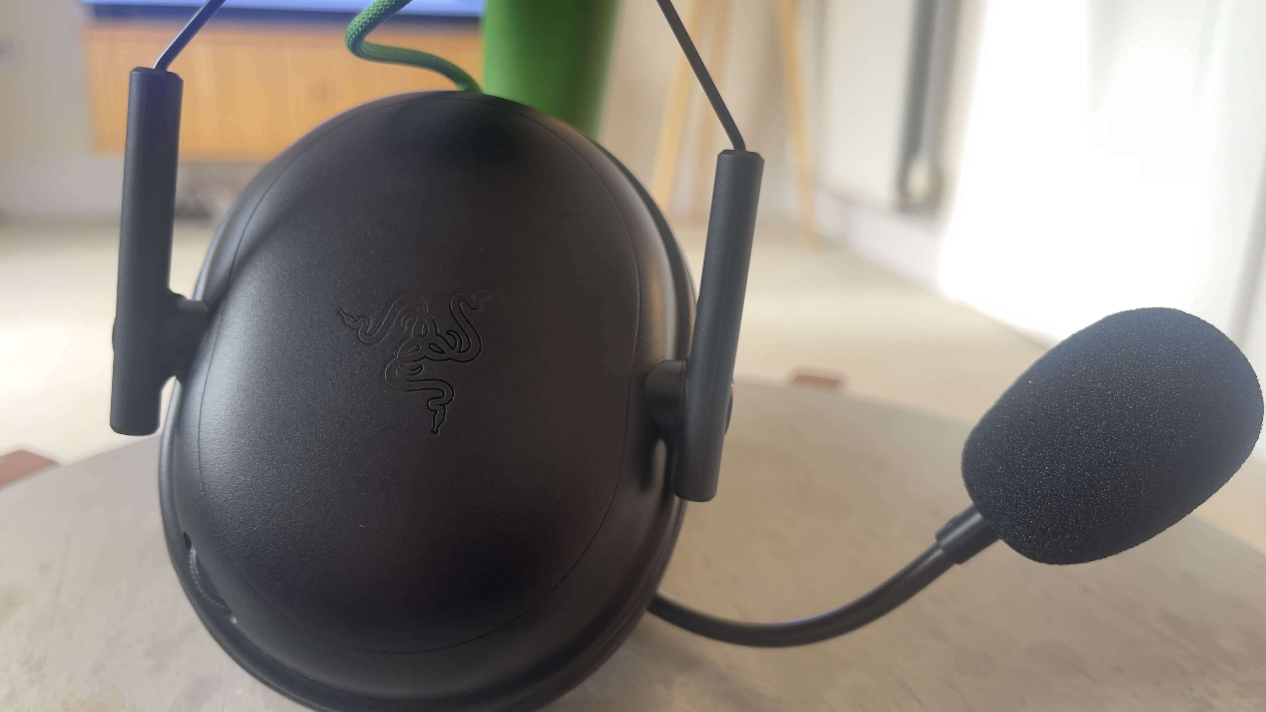 The Razer Blackshark V3 X gaming headset on a table. A living room is visible in the background.