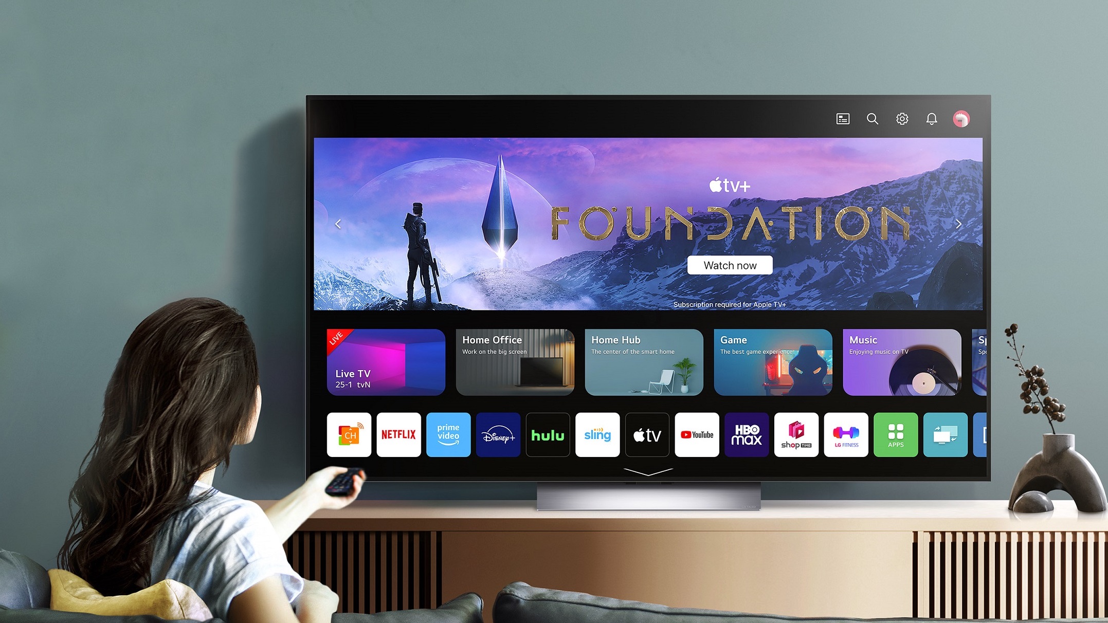 Someone with long brown hair sat in front of an LG C3 OLED TV displaying the home screen against a blue wall