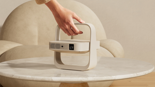 Lifestyle shot of the Xgimi Z6X 5th Generation projector unfolded on a light marble table. A woman's hand is coming from the top of the image to lift the projector up