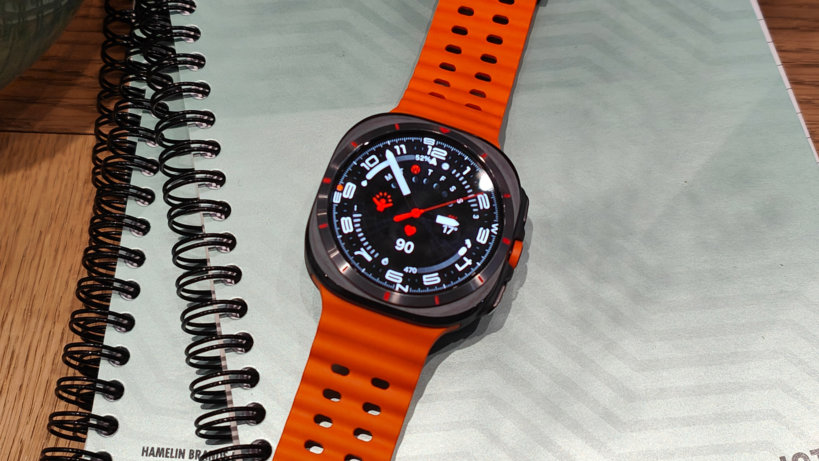 The Samsung Galaxy Watch Ultra pictured on a notebook with an orange strap.