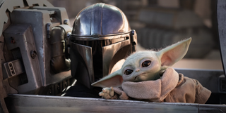 The Mandalorian in full armour including a helmet, with Grogu, a green alien with elf-like ears sitting beside him.