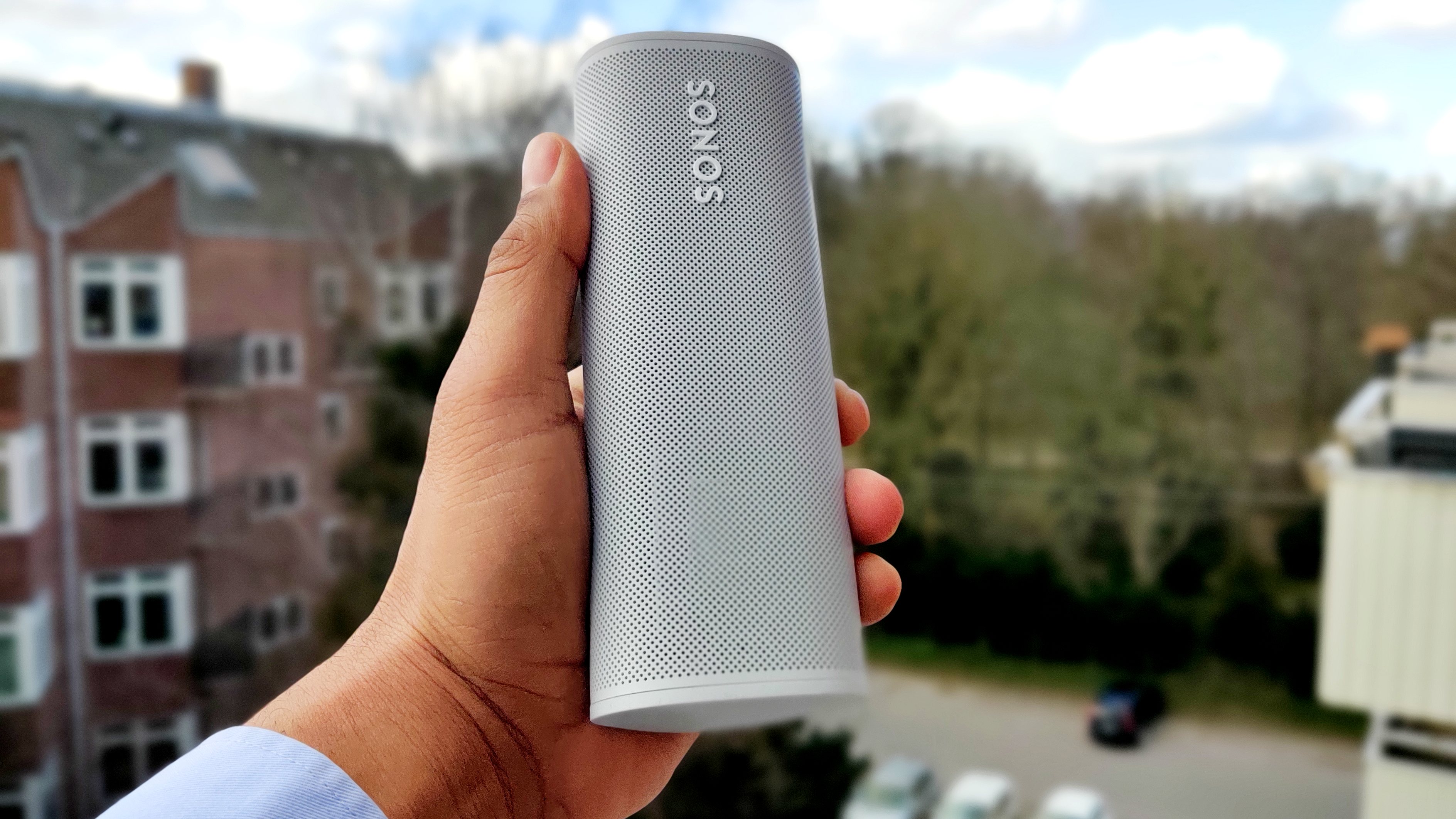 Someone holding the Sonos Roam portable speaker in white up to the camera with a blurry street scene in the background.
