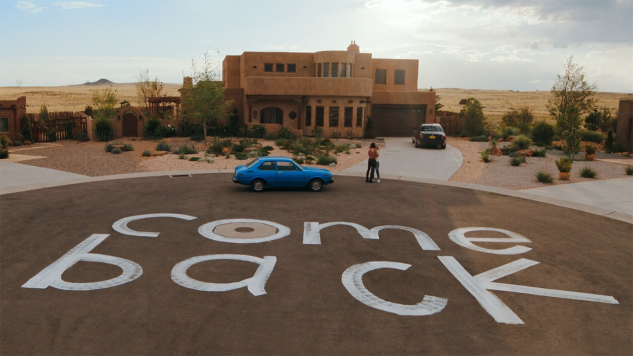 A wide shot of Zosia and Carol hugging near a giant painted 'come back' sign in Pluribus episode 7