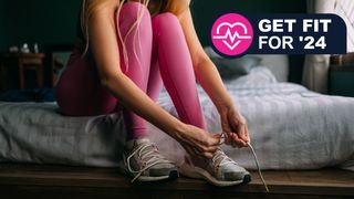 A woman sits on the corner of her bed wearing workout clothes and tying the laces on her running shoes