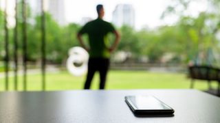 Cell phone was left on an outdoor table with a person's back and a green city in the background