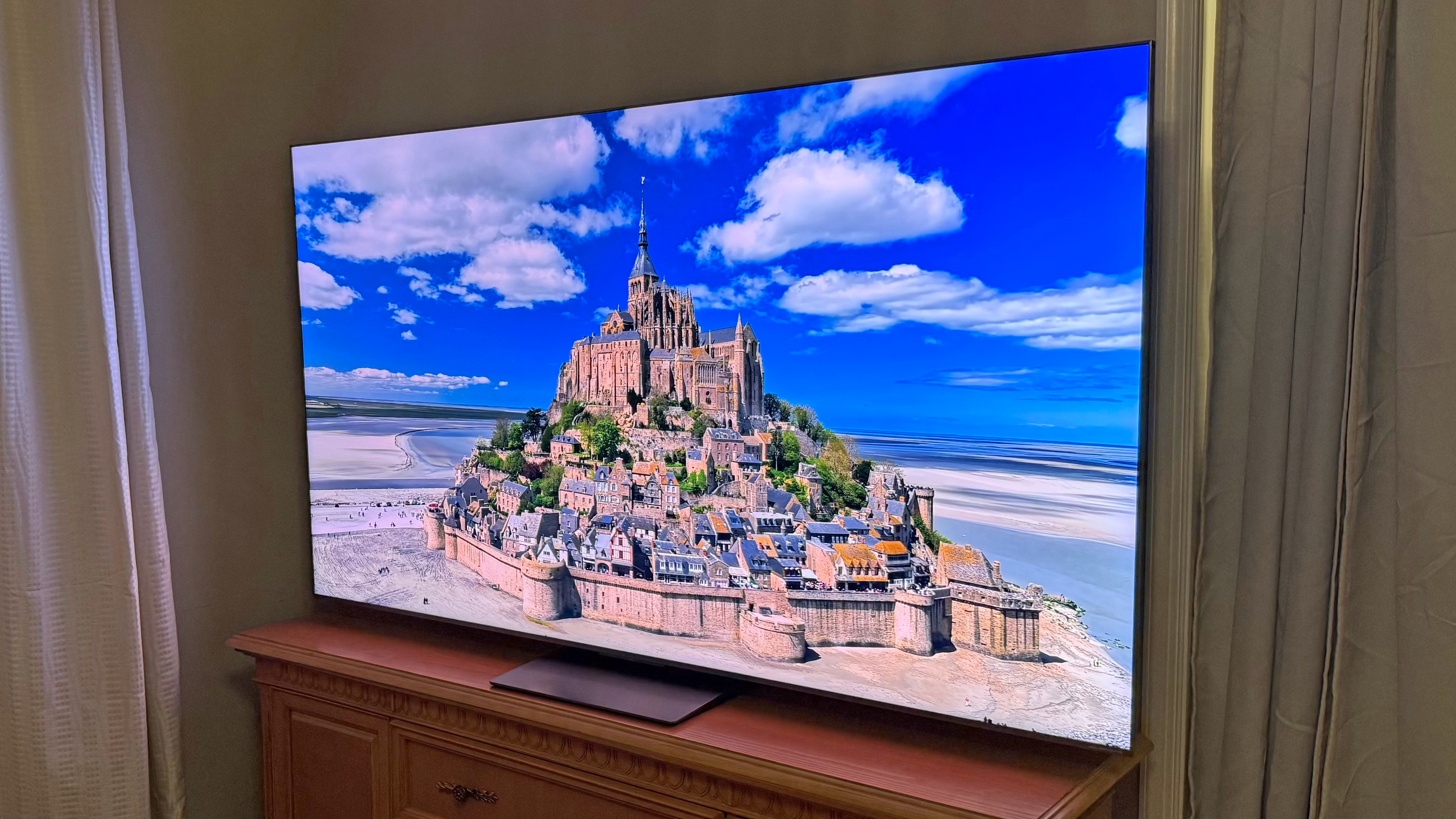 TCL QM8L TV on a wooden TV unit, showing an image of a castle town with a striking blue sky, showing the brightness of the screen