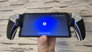 The PlayStation Portal displaying its home screen being in held in a man's left hand in front of a white brick-effect wall