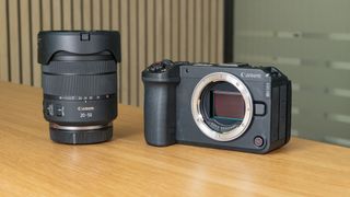 The Canon EOS R6 V mirrorless camera on a wooden table with a pine-slatted backdrop, no lens attached, alongside the RF 20-50mm F/4 L PZ lens