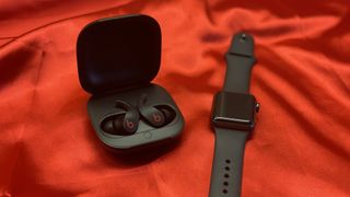 The Beats Fit Pro true wireless earbuds in their charging case on a red backdrop next to an Apple Watch