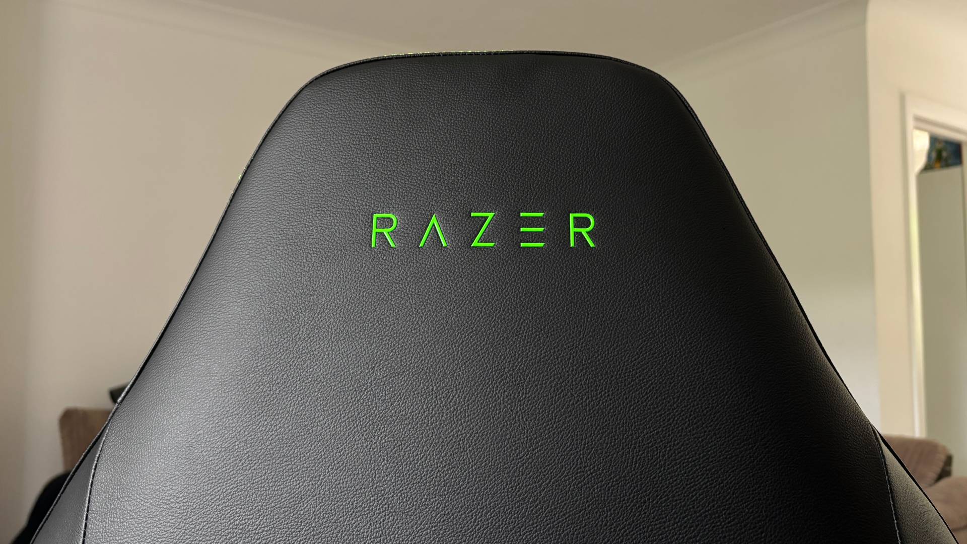 Neon green Razer text logo on a black Razer Iskur V2 NewGen gaming chair with a white wall in the background.