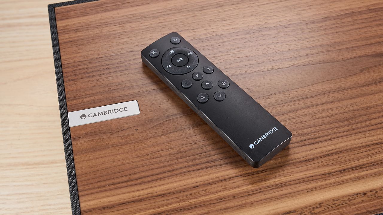 A closeup of the walnut top panelling of the Cambridge Audio Evo One, with its remote control laying on top. The remote control has buttons for power, mute, playback, input switching, and brightness control for the speaker's screen.