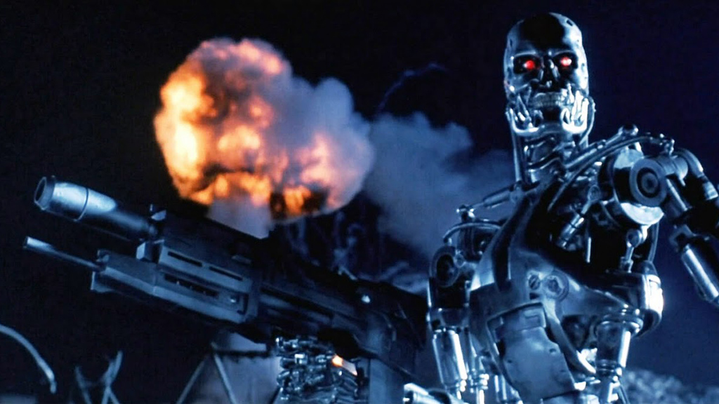 A still from Terminator 2 in which a Terminator model holds a big gun and there's an explosion in the background.