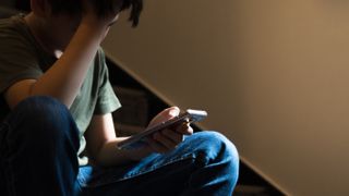 Young Asian preteenteenage boy sit on stair at home, cry and cover his face with hand, other hand hold smartphone, feeling alone, stressed, frustrated, overwhelmed, depressed. Cyber bullying concept.