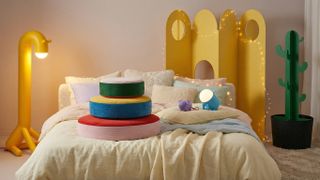 IKEA Grejsimojs homeware products including a dog-shaped lamp, giraffe lamp, cactus coat hook and floor cushions on a bed, with twinkle lights in the background