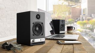 Audioengine HD3 Next-Gen (2025) speakers on a wooden desk next to a phone and laptop and set of keys. There's a window at the back and you can see a nice garden.