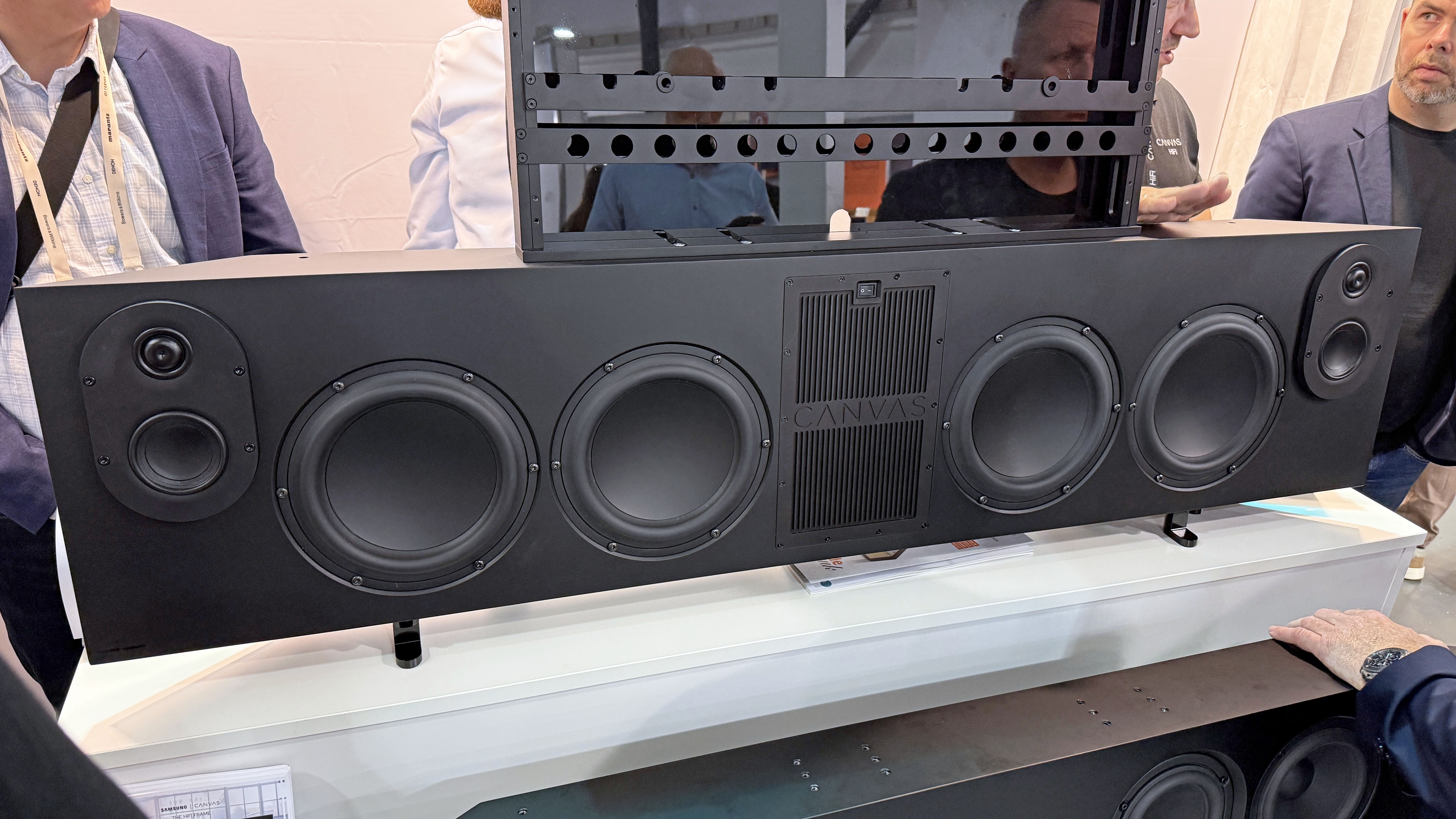 The Canvas L soundbar on a stand at a convention. The grille has been removed, so you can see the speaker arrangement and the mounting bracket