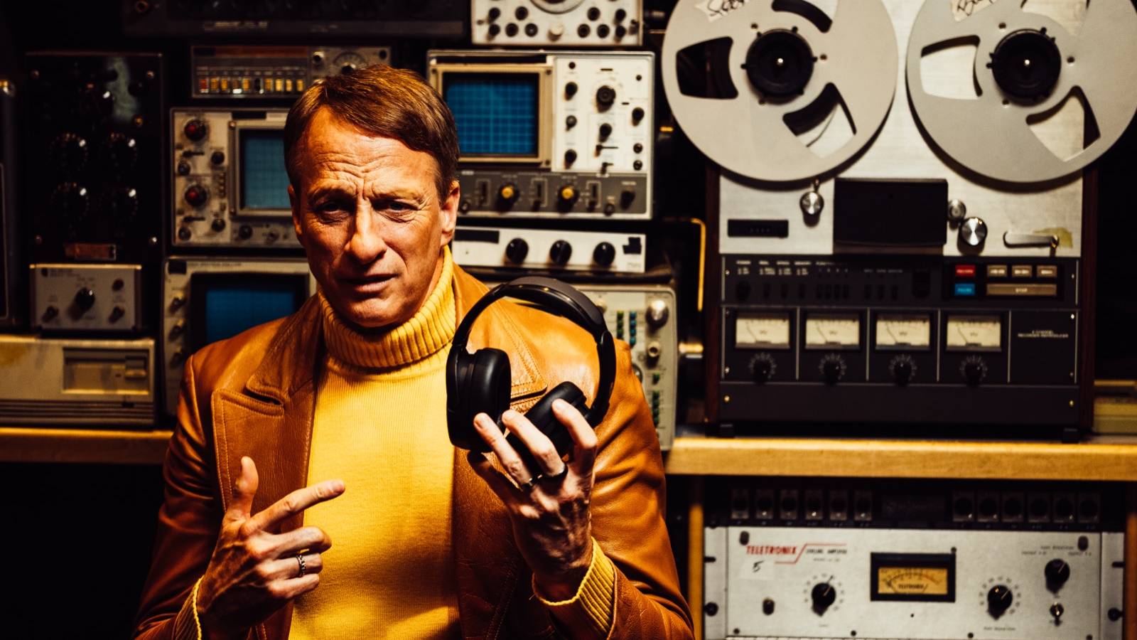 Tony Hawk holding a pair of Skullcandy Crusher 1080, in front of some tape recording machines.