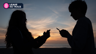 A teenage girl and a teenage boy look at an iPhone smartphone screen on a beach as the sun sets + TechRadar's Leave No Trace logo on the upper left