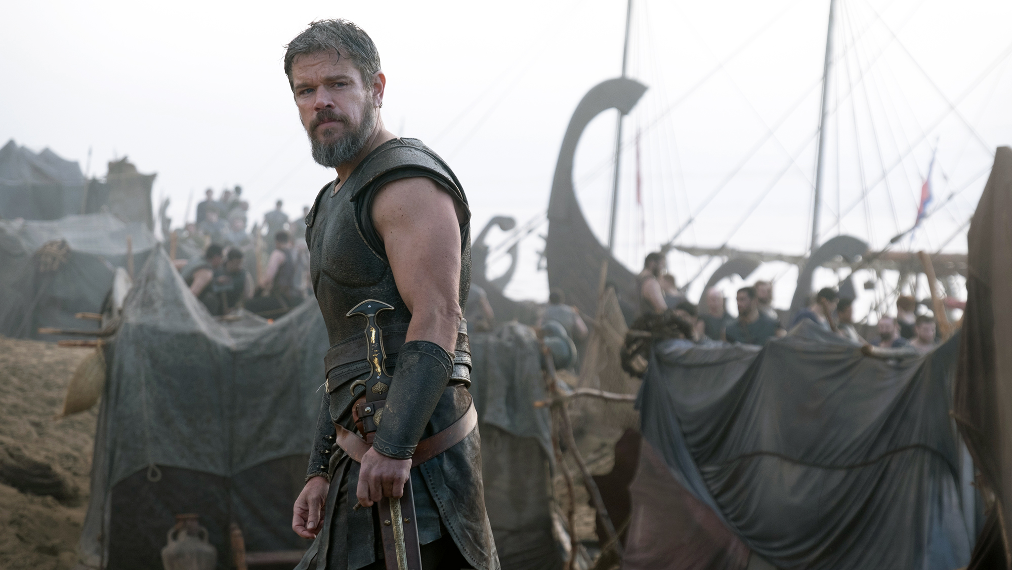 Matt Damon's Odysseus looking over his left shoulder with some ships in the background in The Odyssey