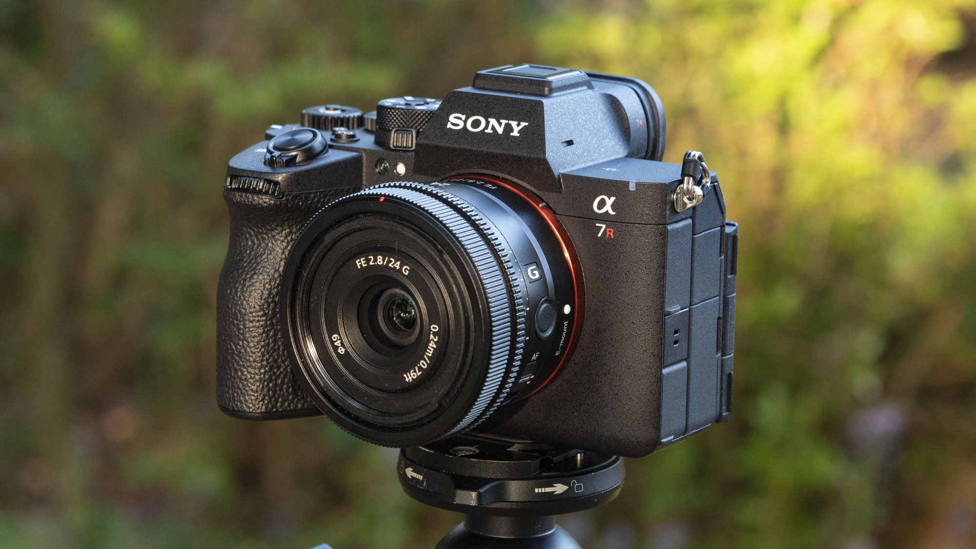 The Sony A7R VI with 24mm f/2.8 lens attached, mounted to a tripod outdoors against a background of out-of-focus green foliage