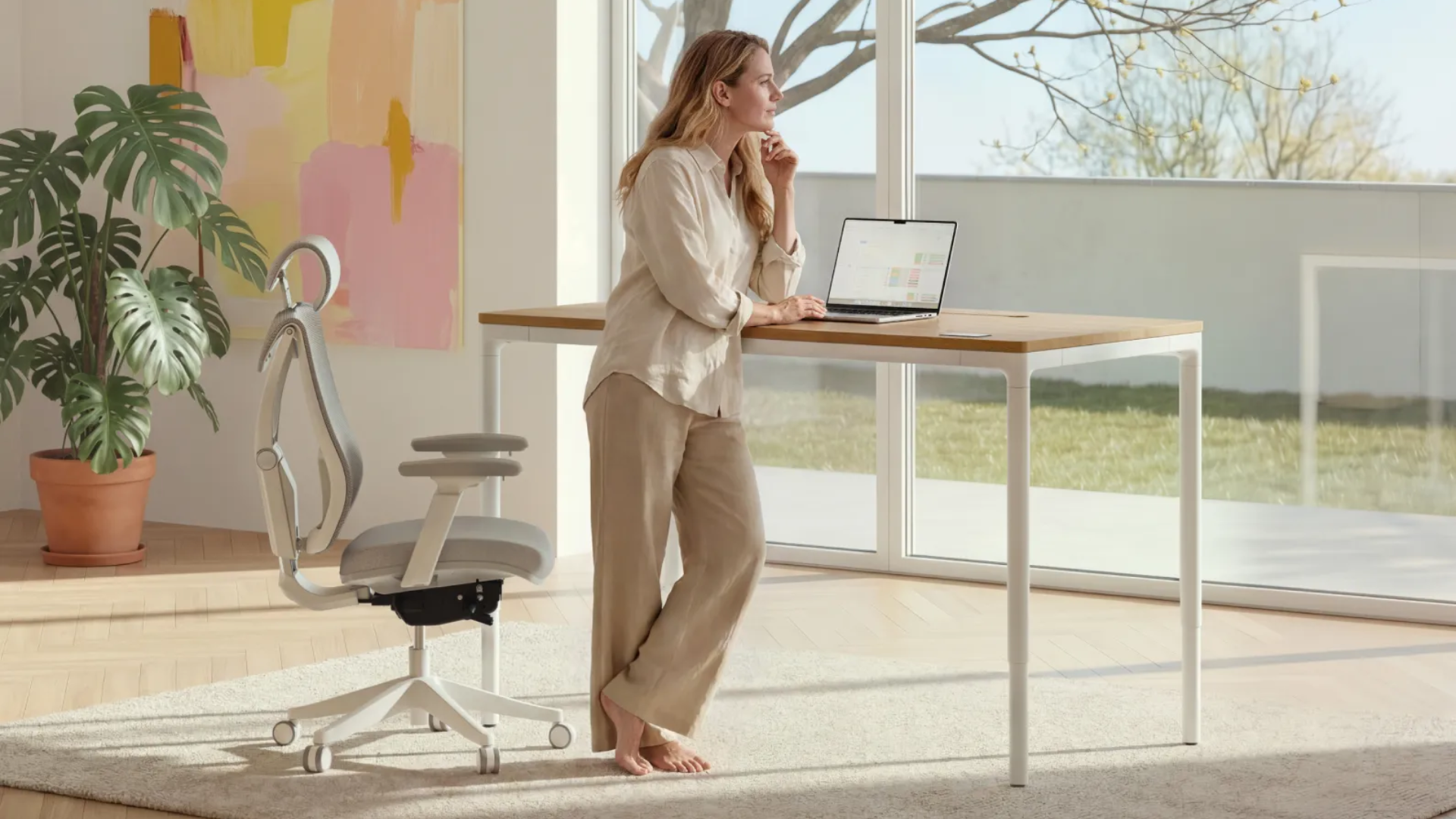 A woman with a laptop standing at the Desktronic HomeMax desk with a SitPro office chair behind her