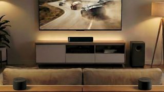 Majority Bowfell Halo Atmos in a neutrally decorated room, shot from behind the sofa. Two satellite speakers are in the foreground and there's a TV on the wall with the soundbar below it and subwoofer to one side of it