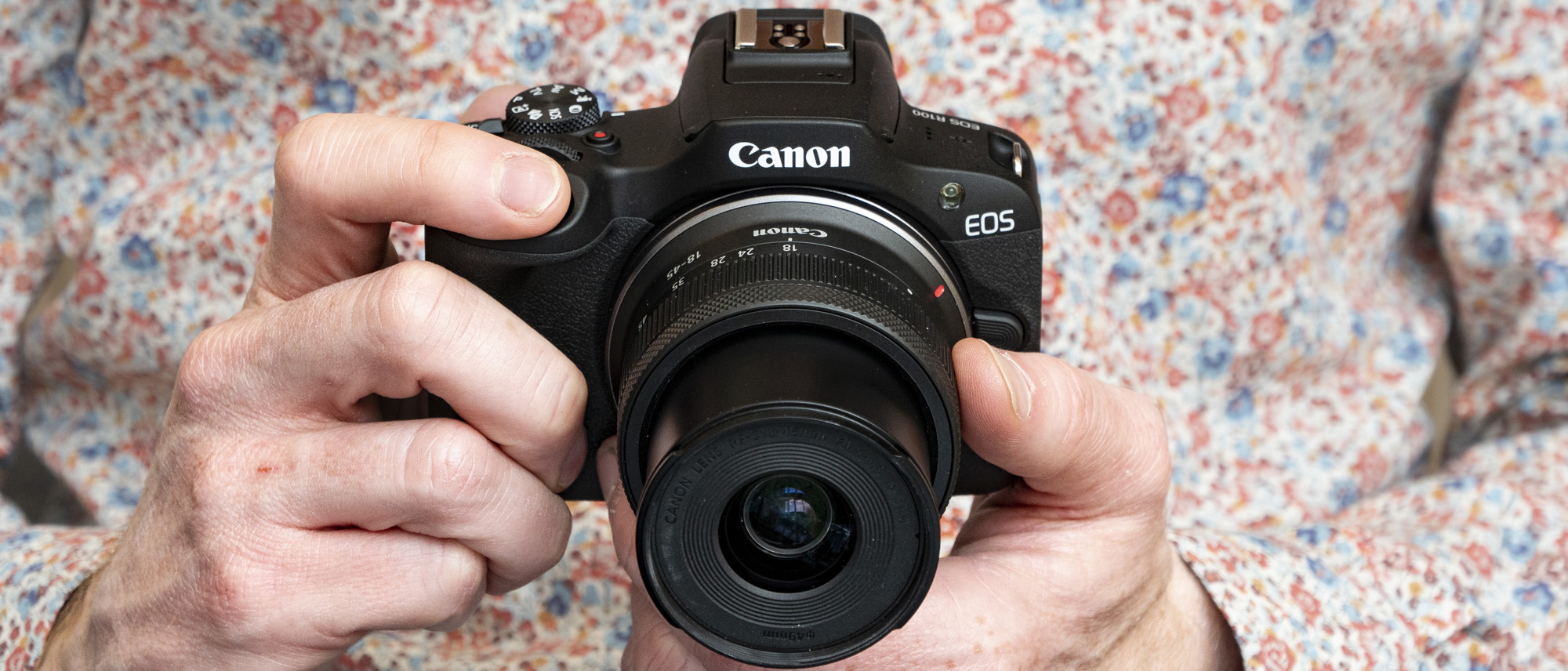 Canon EOS R100 camera in the hand, with person's flower-print shirt in background