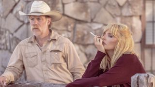 Beth smokes while leaning against a wooden fence with Zachariah standing behind