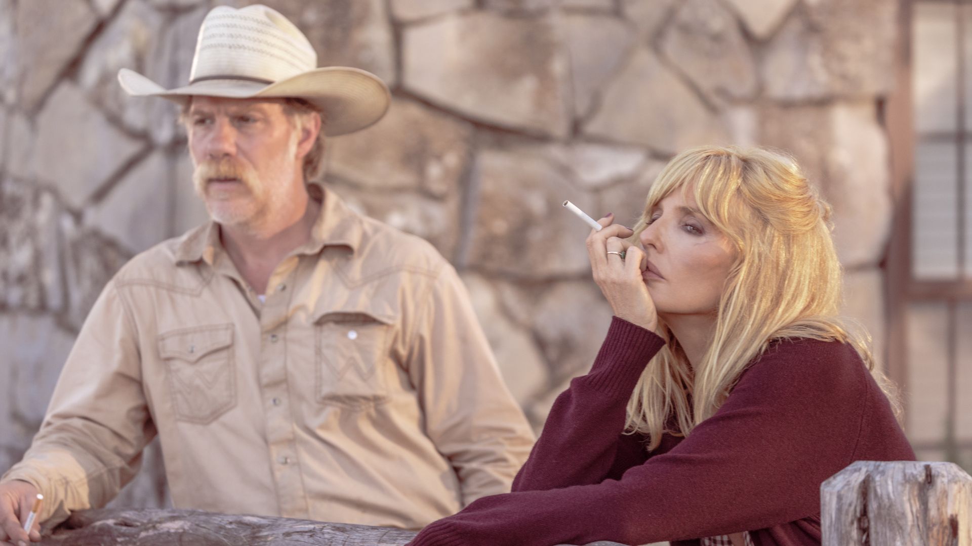 Beth smokes while leaning against a wooden fence with Zachariah standing behind