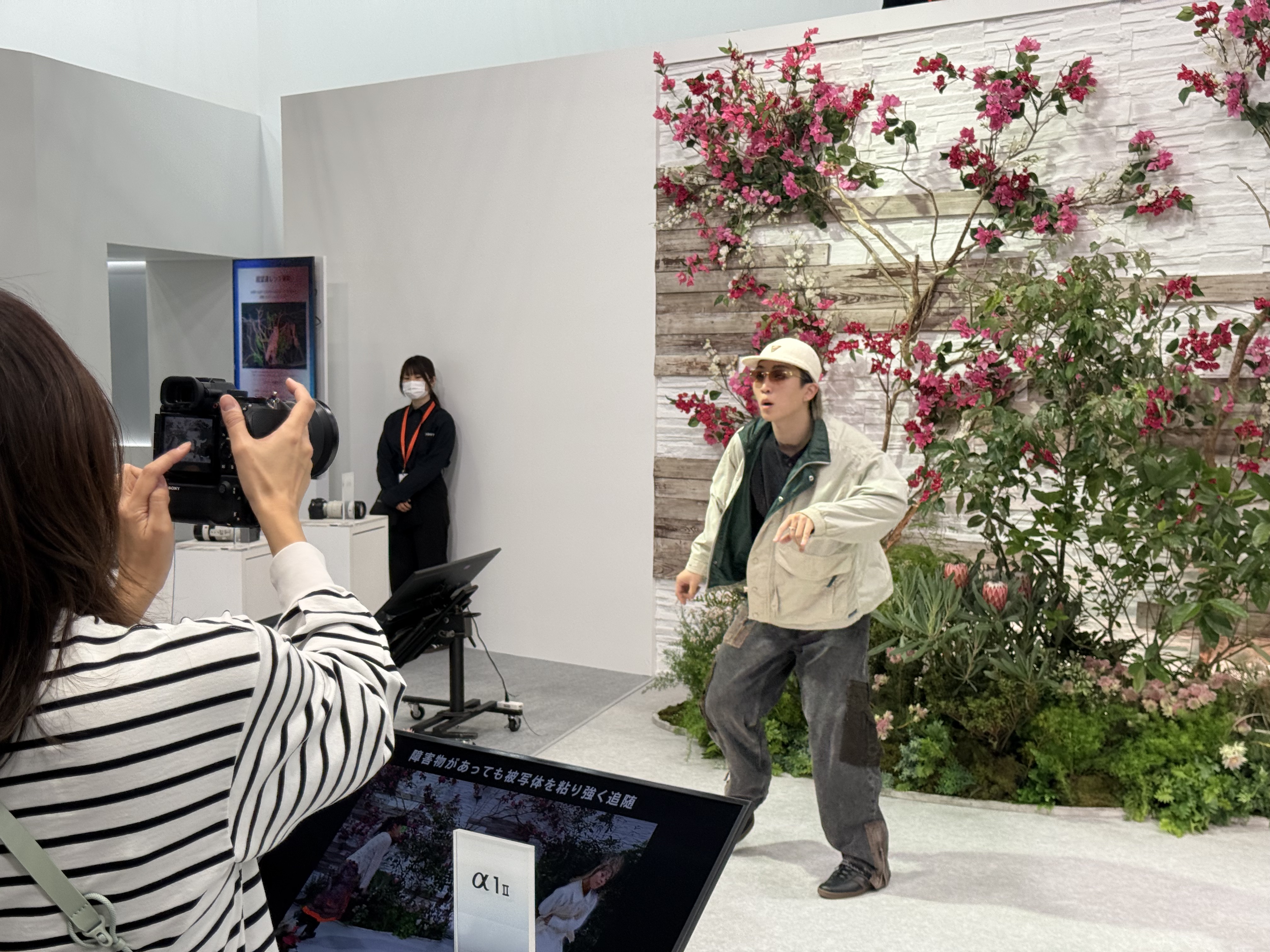 A dancer in front of a floral background, being photographed by a CP+ 2026 show visitor with a Sony camera, at the Sony stand
