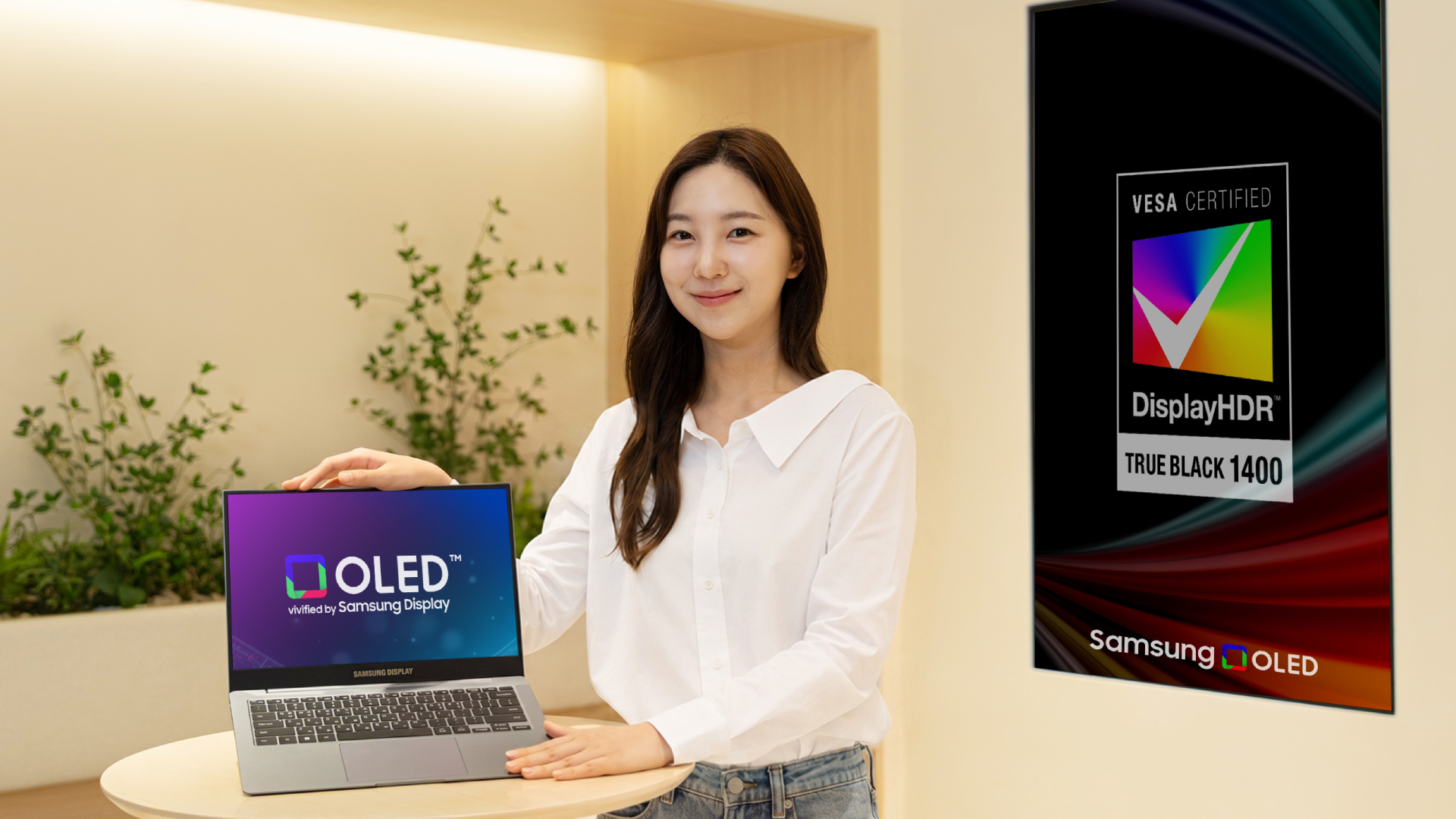 A young woman holding a laptop with OLED written on the screen. A poster can be seen behind with DisplayHDR True Black 1400 written on it behind her