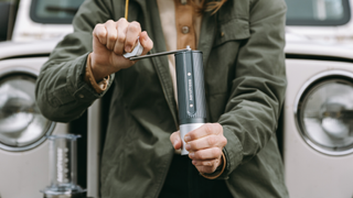 Lifestyle image of the AeroPress Manual Coffee Grinder being used by a person in a dark green coat in front of a vintage white car
