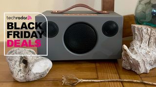Audio Pro Addon T3+ on a table, with kind of witchy vibes (a seaglass-filled vase to one side, a hagstone to the other and a poppy-seed case underneath it) and TR's Black Friday deals badge in pink