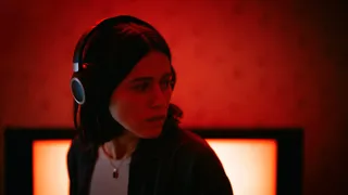 Nina Kiri as Evy in Undertone, surrounded by a red room. She is wearing headphones and looks nervously at something. 