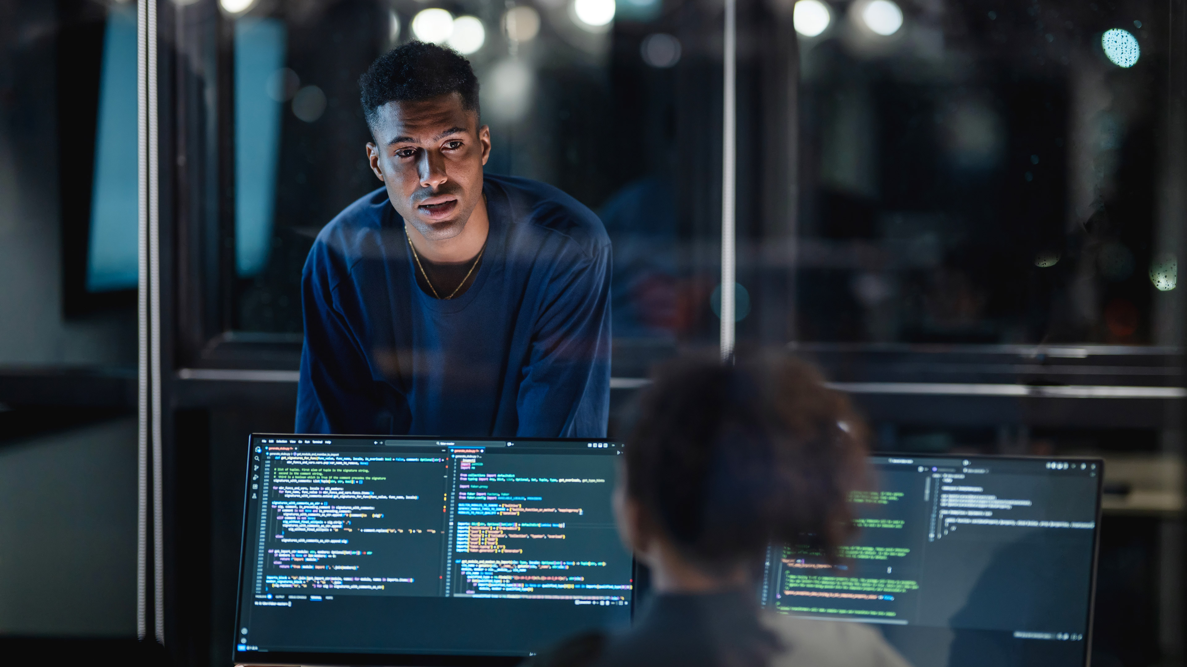 An office worker speaking to a colleague in an office at night. The co-worker is seated in front of computers
