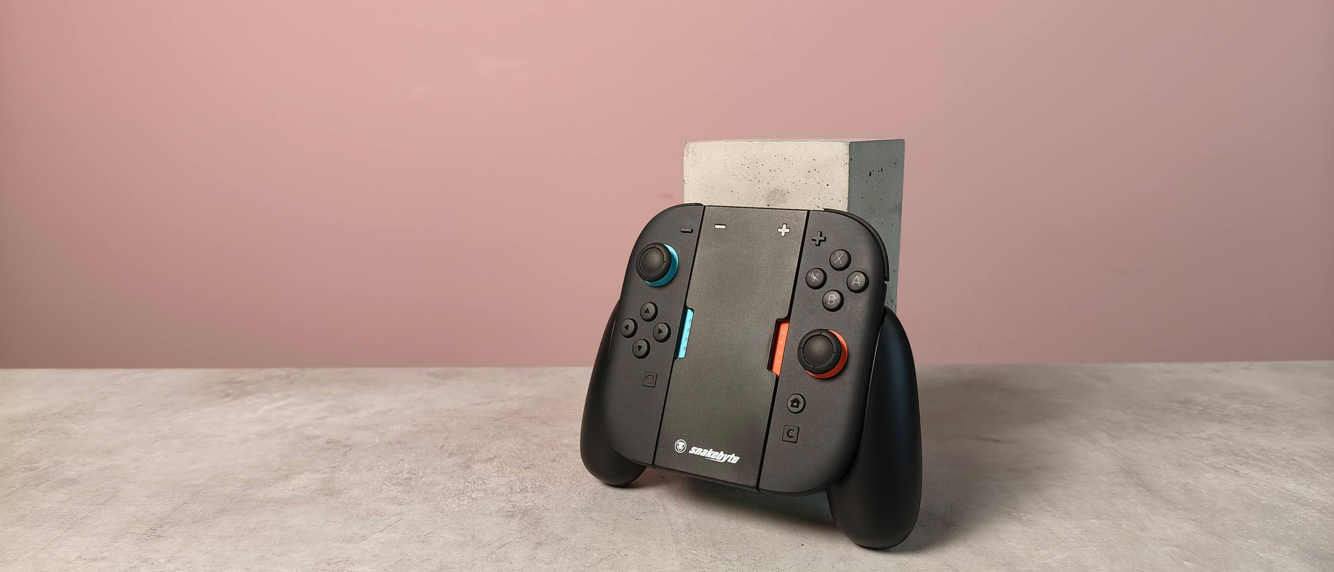 Snakebyte Ergo Grip S2 with Joy-Con 2 controllers attached, leaning on a plinth on a desk with pink wall in background