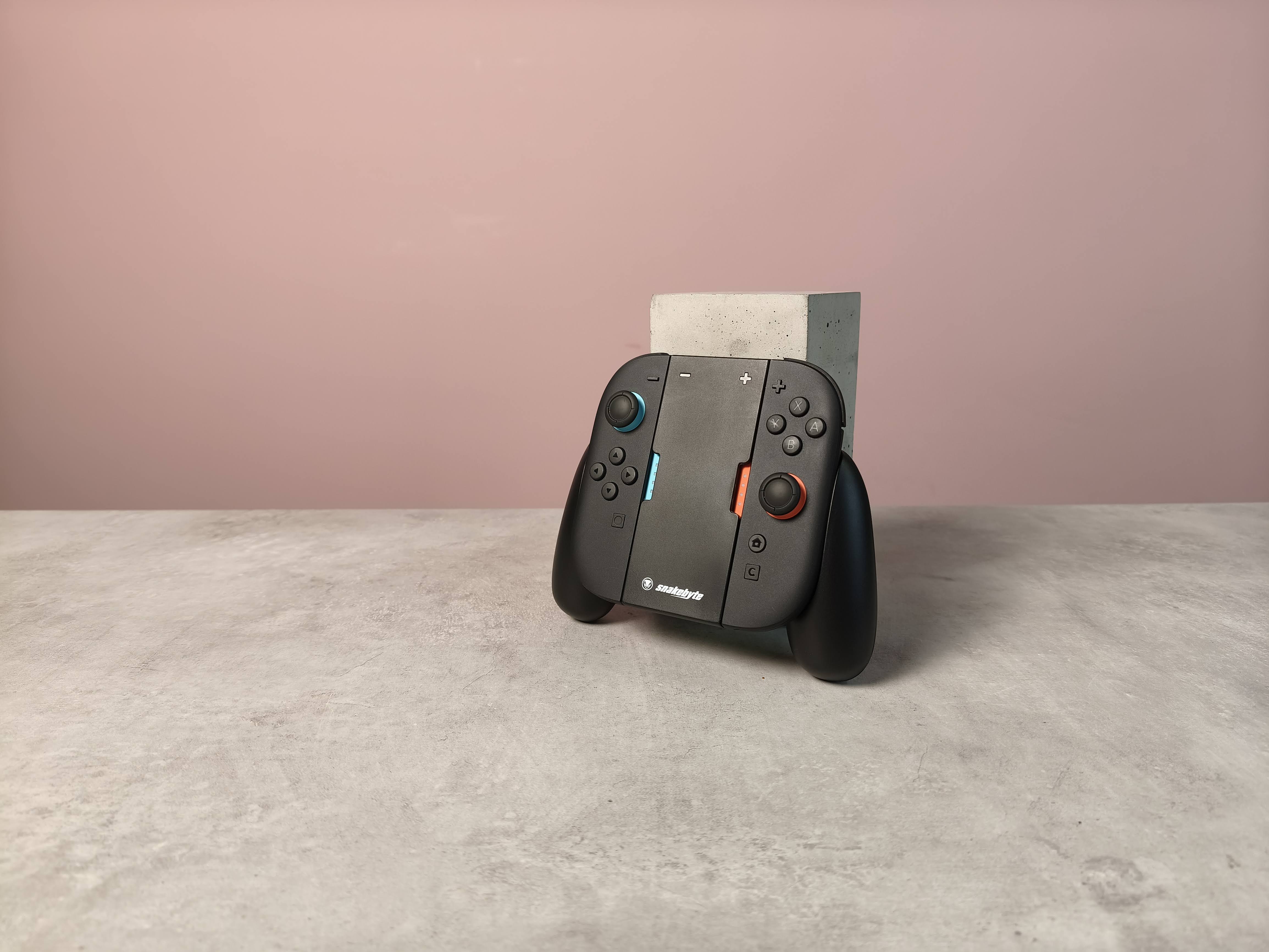Snakebyte Ergo Grip S2 with Joy-Con 2 controllers attached, leaning on a plinth on a desk with pink wall in background