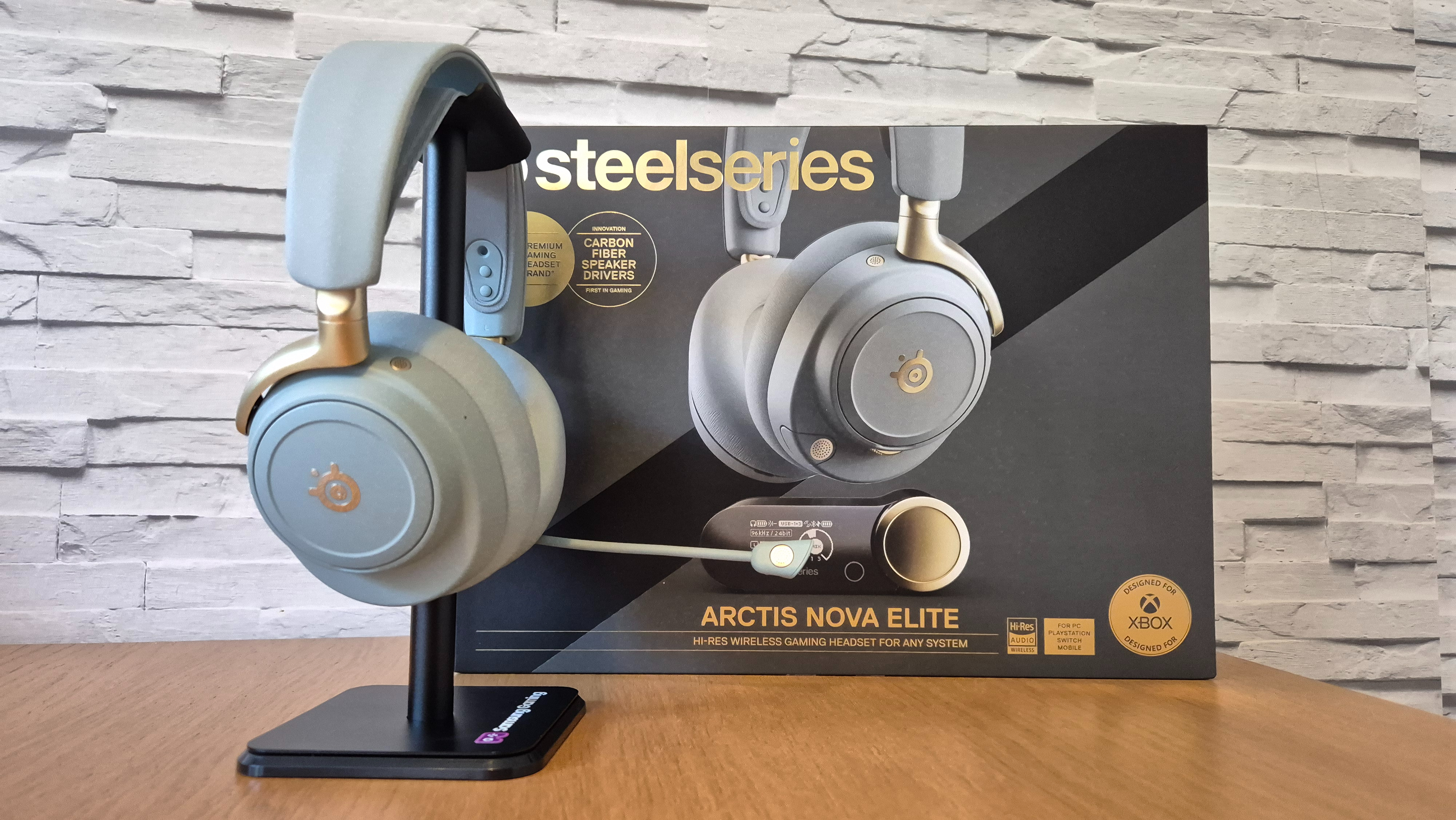 The SteelSeries Arctis Nova Elite headset in front of a white brick background and on a wooden surface
