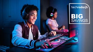 Two teenage gamers sitting side-by-side at a large desk while wearing headphones, using a mouse and keyboard and each looking at a bright monitor that illuminates their faces in a darkened room.