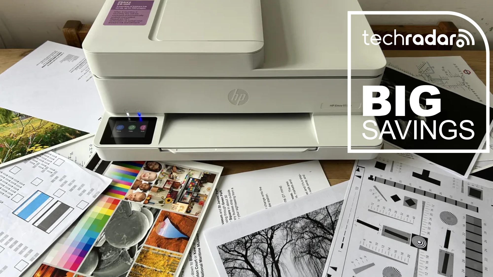 A HP Envy 6555e printer during our review, with freshly printed paper scattered around, and a TechRadar badge that reads 'Big Savings'