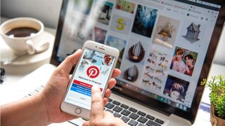 Pinterest app on a smart phone with a laptop behind and coffee on the desk