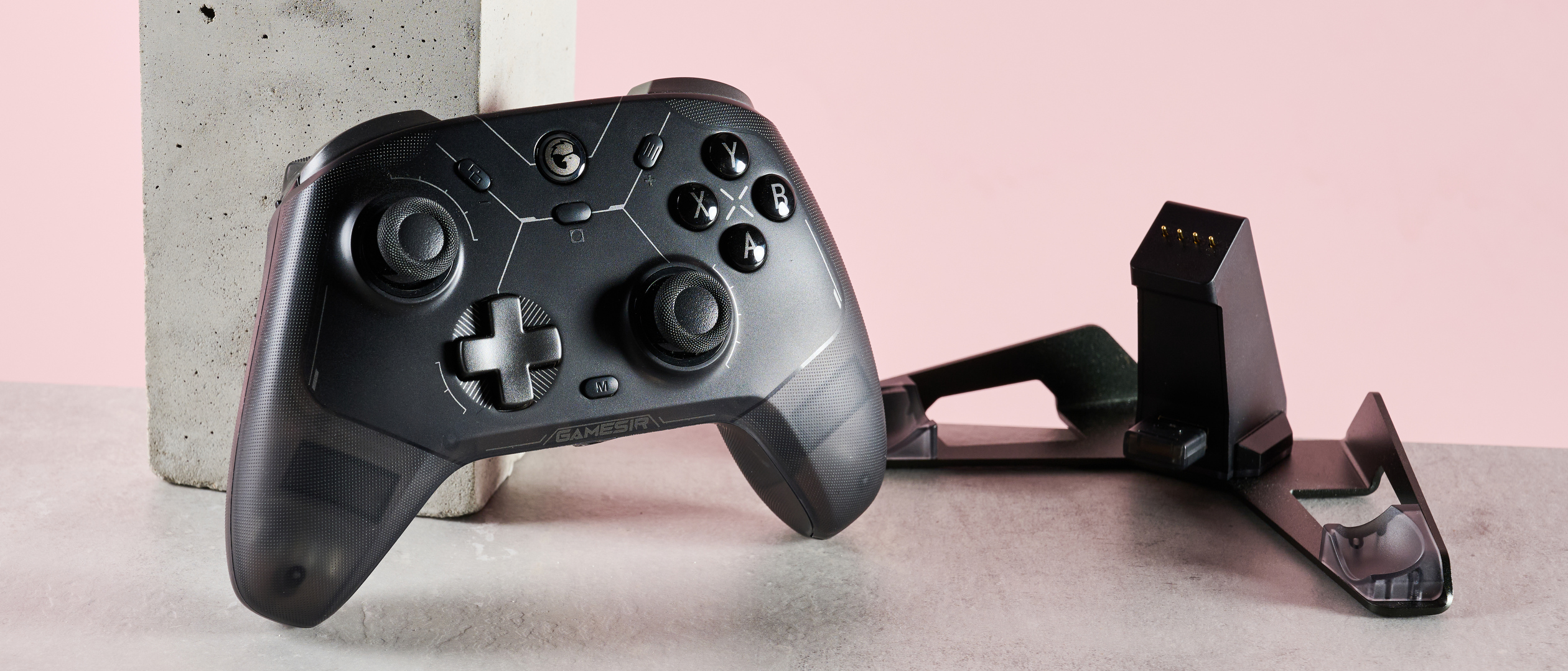 GameSir Cyclone 2 leaning on plinth next to charging stand, on desk with pink background