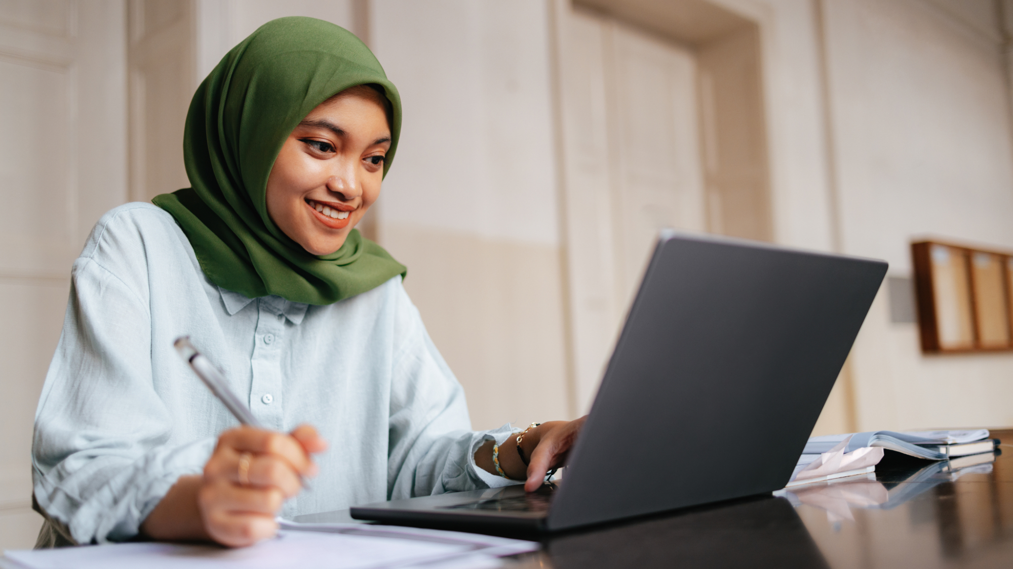 Indonesian student wearing hijab, sitting and using a laptop computer. She is at the university doing homework or assignment. She is smiling and looking down at the screen while typing on the keyboard.