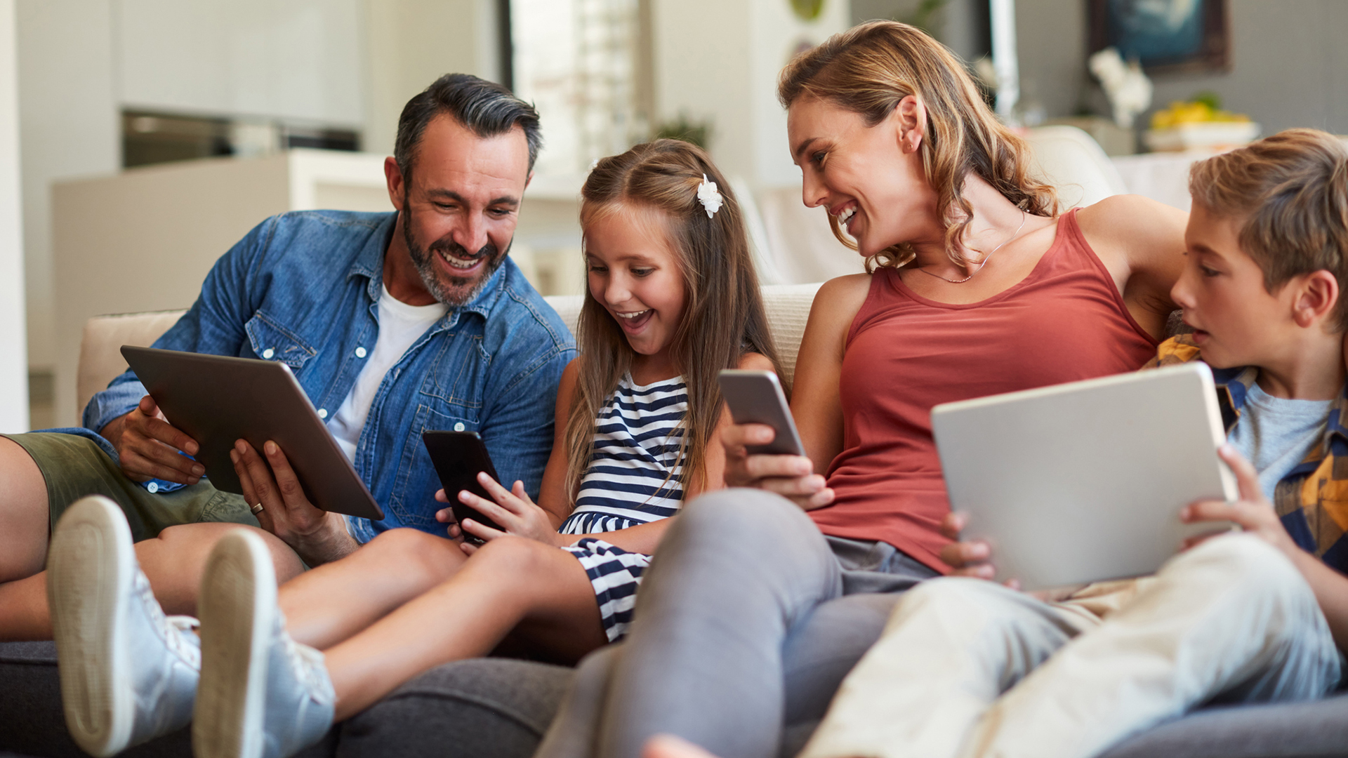 A family sits together on a couch playing games on their phones and tablets