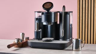 The Meraki Espresso Machine with a tamp, coffee distributor and portafilter to its left and a milk jug to its right, in front of a pink and wooden effect background.